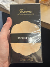 Load image into Gallery viewer, A hand holding a boxed Rochas Femme Collection Haute Parfumerie perfume spray. The box is black with gold lettering and a large beige flower-like graphic on the front.
